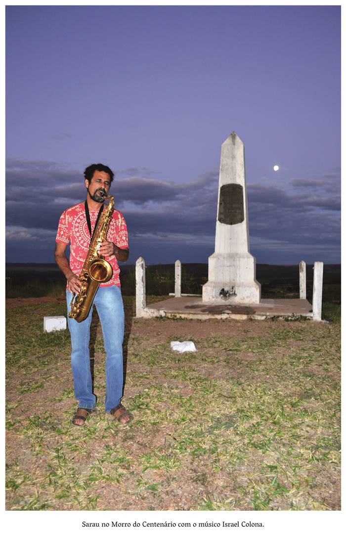 4 z 7: Sarau no Morro do Centenário com o músico Israel Colona. A imagem retrata o anoitecer na Pedra Fundamental que está ao fundo da fotografia colorida e a lua cheia entre nuvens embelezam a paisagem. Um músico vestido de camiseta com estampa vermelha e branca, calça jeans e sandália de couro, toca saxofone de cor dourada. O músico é pardo, tem cabelos e barba preta. Realização: Academia Planaltinense de Letras, Artes e Ciências (APLAC), pelo Ecomuseu Pedra Fundamental e pelo Coletivo Nativo Audiodescrição produzida  pelo Instituto de Promoção das Pessoas com Deficiência Visual Audiodescritora: Elma Lúcia Rodrigues Consultor: Fernando Rodrigues Este projeto é promovido com recursos do Fundo de Apoio a Cultura do DF